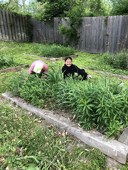 a woman in pink is weeding a garden bed while a young girl in black smiles while sitting in the garden bed.