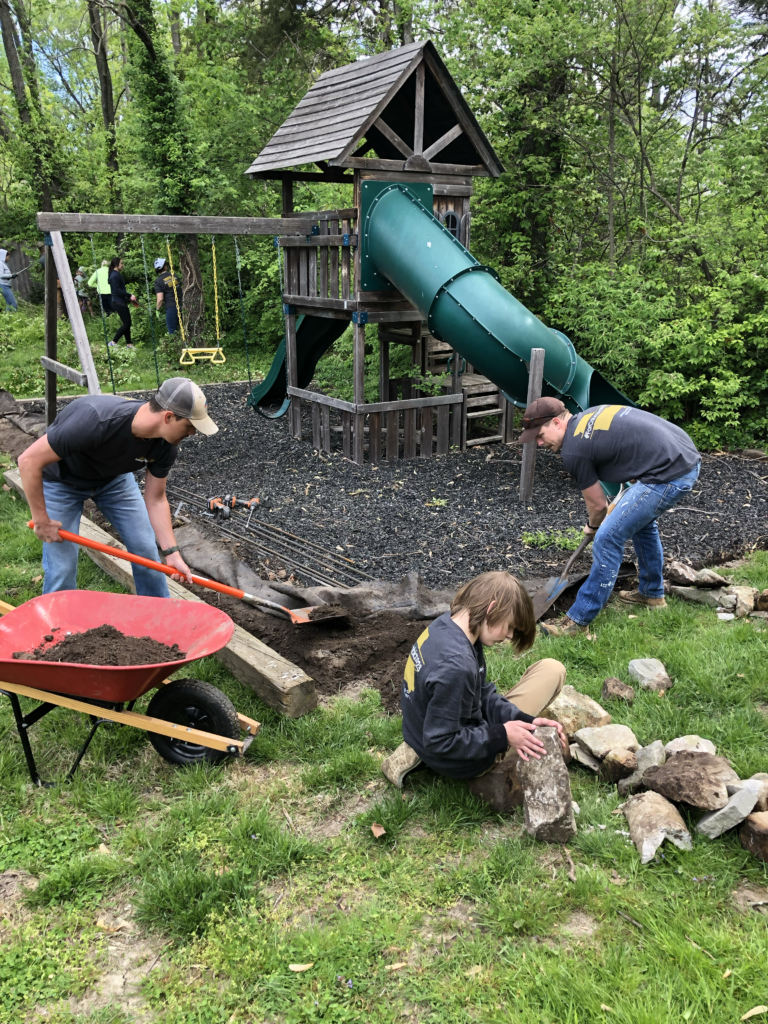 Two men are digging near a playground while a young boy moves rocks for a walkway